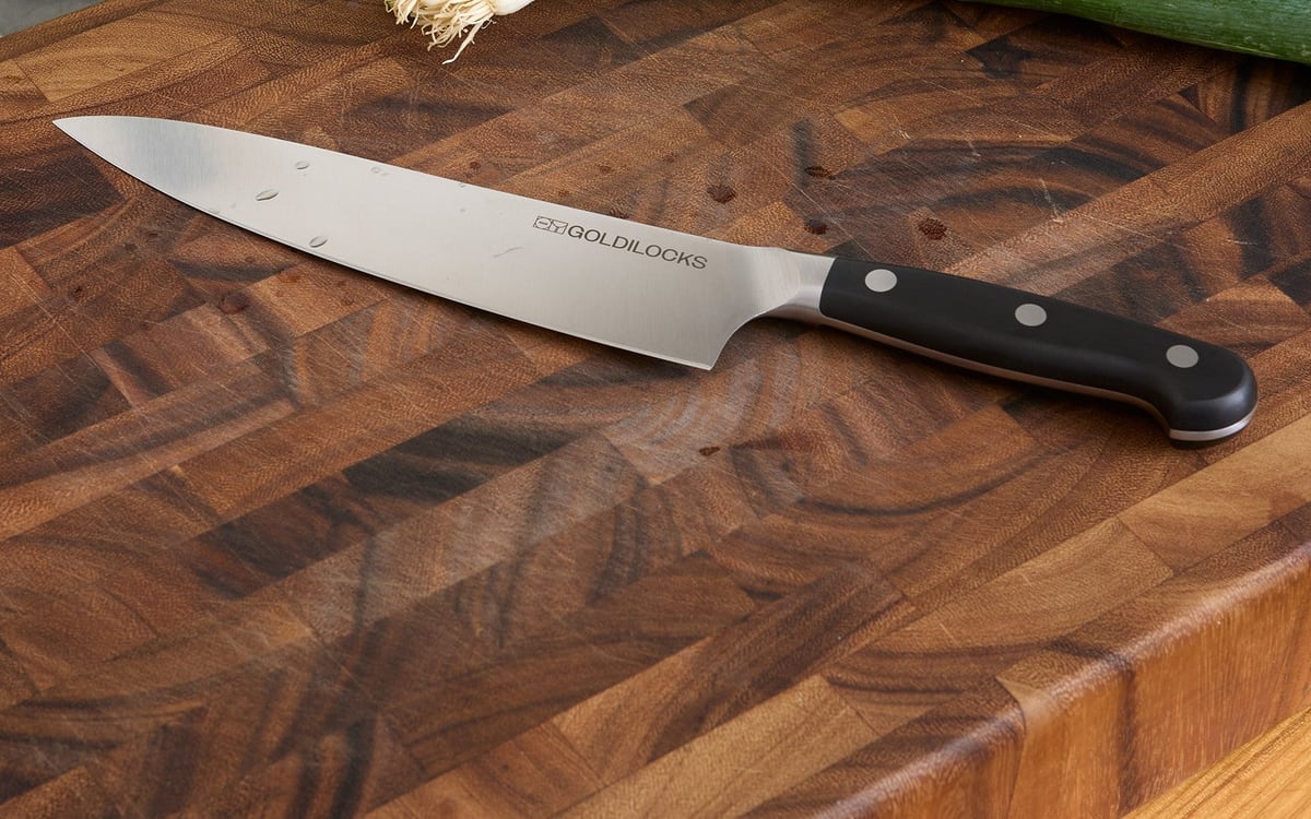 Goldilocks chef’s knife lying on a dark end-grain wooden cutting board, tip to the left, black riveted handle