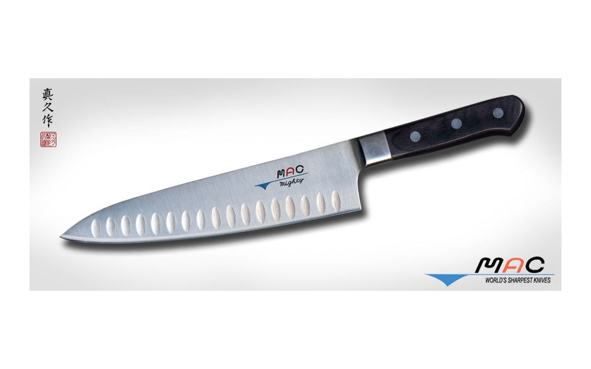 MAC MTH-80 chef’s knife on a pale grey ground: a stainless blade with a row of oval dimples, tip to the left, and a black riveted handle