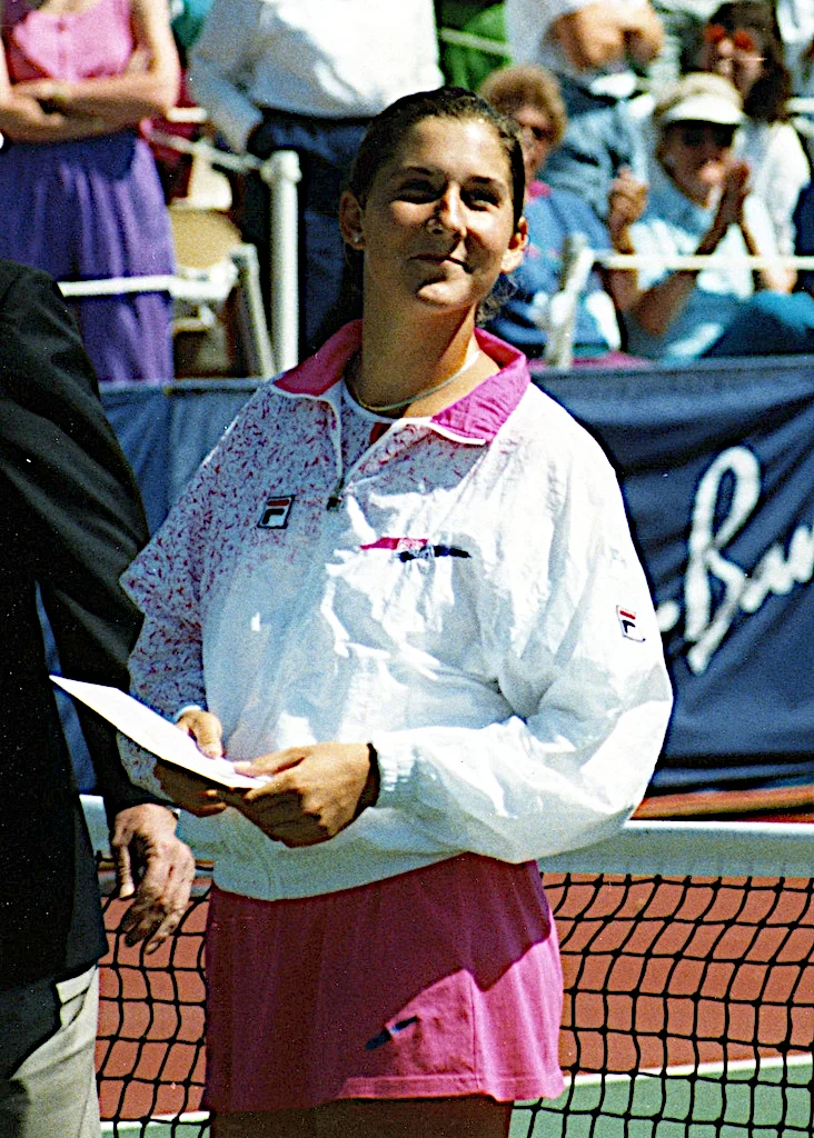 Color photograph of Monica Seles holding a presentation envelope by a tennis net, in a white and pink FILA jacket and skirt.