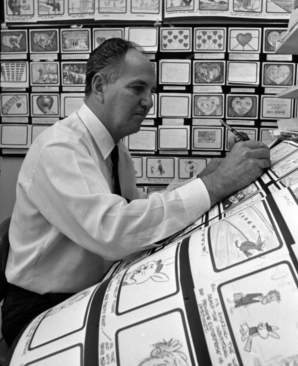 Carl Urbano draws at a sloping desk with rows of storyboard panels behind him.
