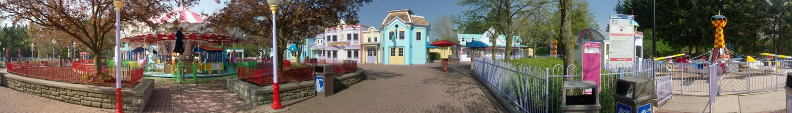 Wide panorama of a colorful theme-park street with a carousel, pastel facades and small rides.