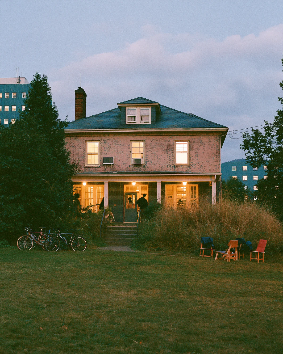 A weathered shared college house glowing at blue hour as students arrive by bicycle